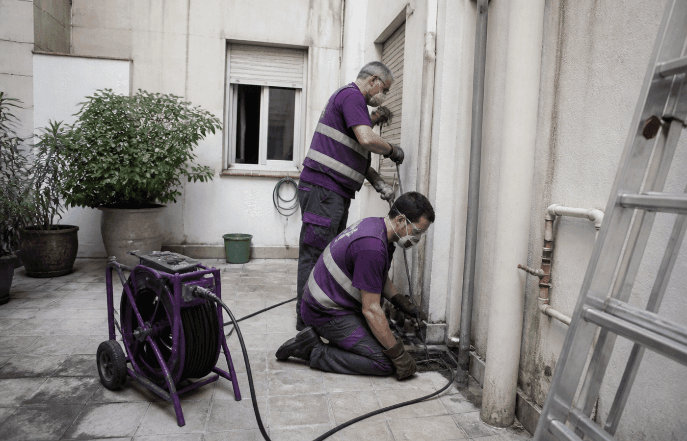 Dos técnicos trabajan en el patio comunitario revisando una arqueta junto a la bajante de un bloque residencial